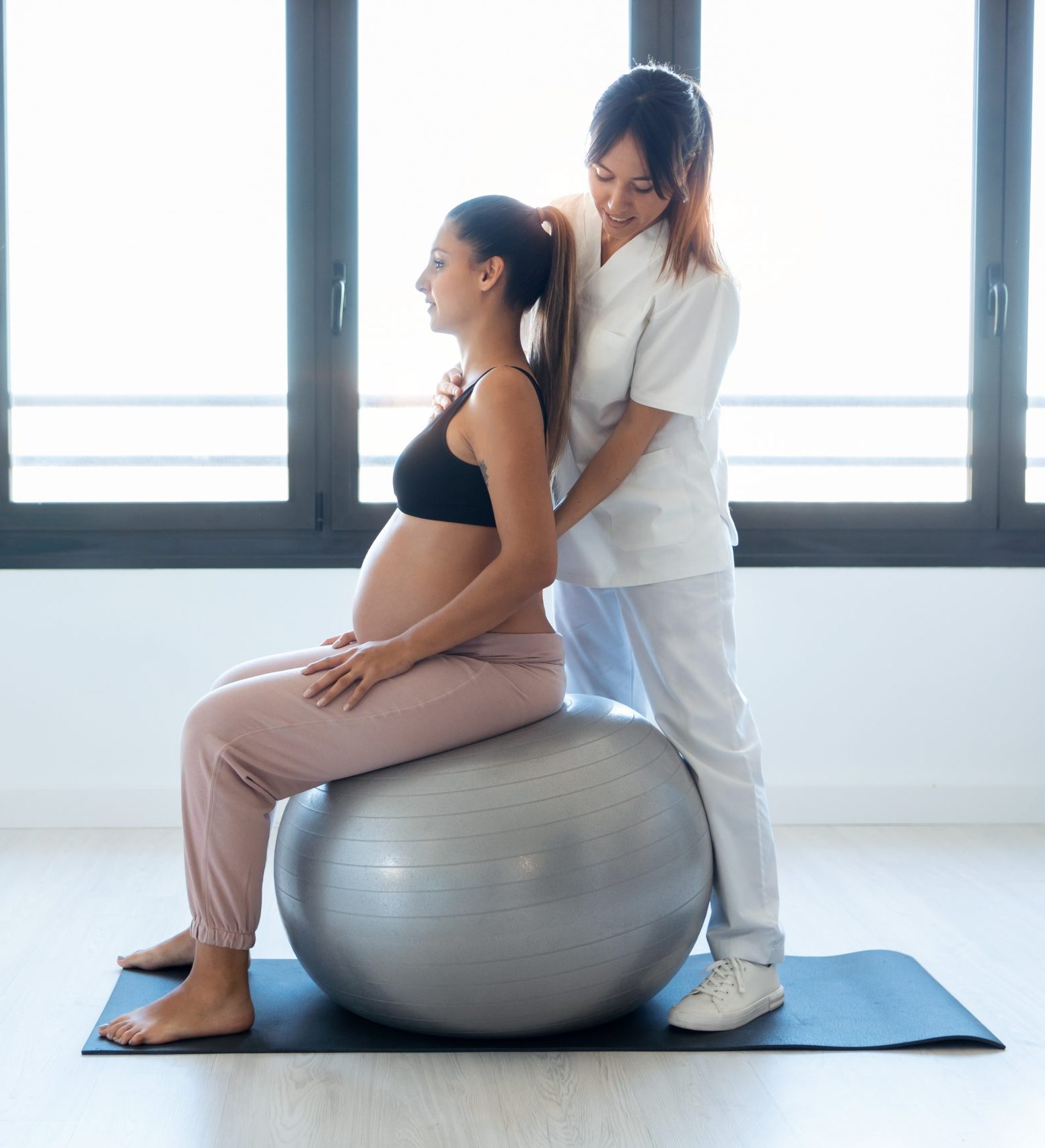 Beautiful young physiotherapist helping to beautiful pregnant woman for doing pilates exercises with ball preparing for childbirth at home.